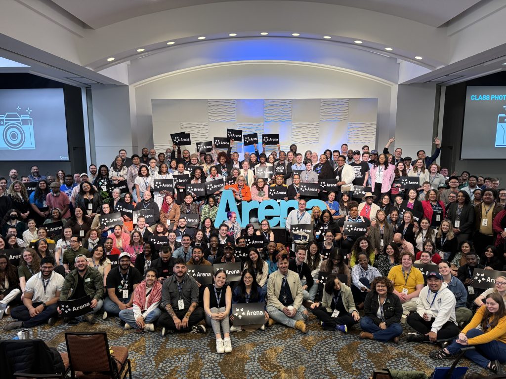 Group photo of hundreds of people surrounding large cut out letters that say Arena holding placards that say Arena, smiling.
